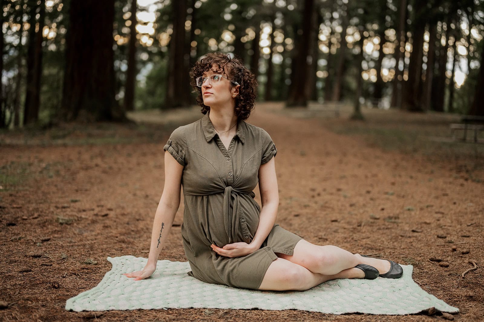 Pregnant woman in glasses sits cross-legged on a blanket in a forest at Mt Tabor during her Maternity photo session, looking to the side with a gentle expression, hands resting on her belly as soft sunlight filters through the trees.