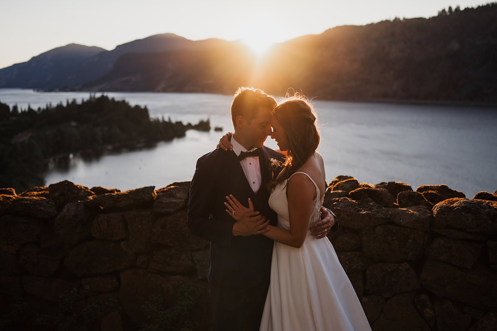 A bride and groom embrace at sunset beside a stone wall, with a the Columbia River and mountains in the background during their micro wedding at the Griffin House.