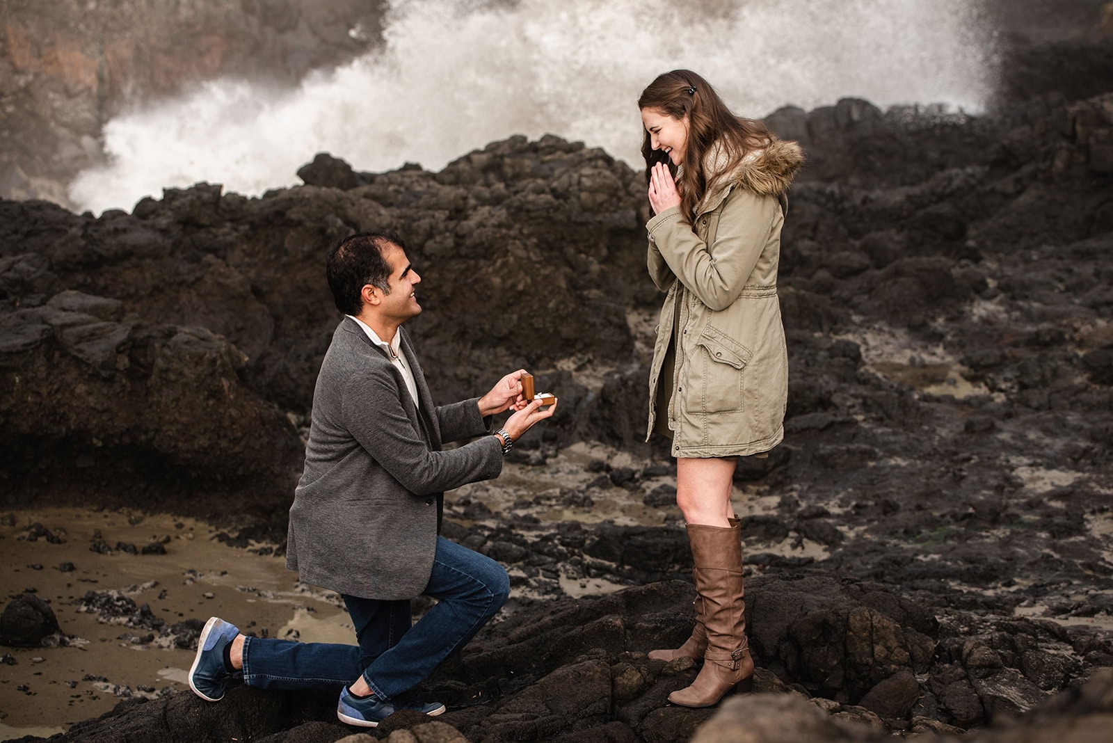 A man kneels on rocky terrain while he proposals on the Oregon Coast near the ocean, holding out a ring to a woman who appears surprised and emotional, with waves crashing against rocks in the background under a cloudy sky.