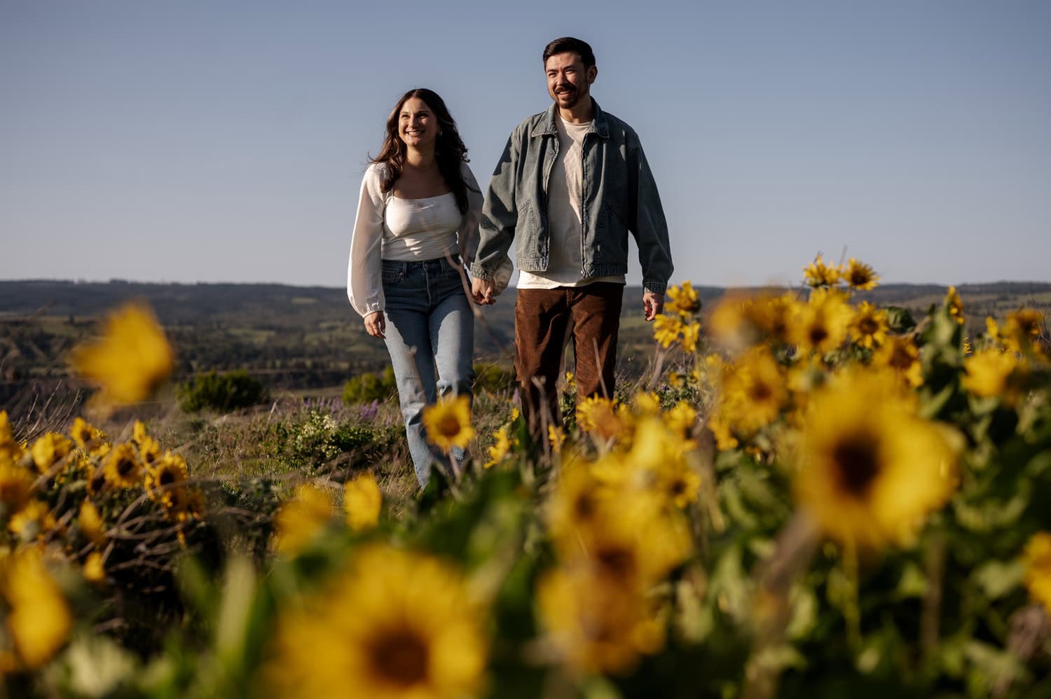 A couple holding hands walks through a field of wildflowers at Rowena Crest under a clear sky during their engagement session.