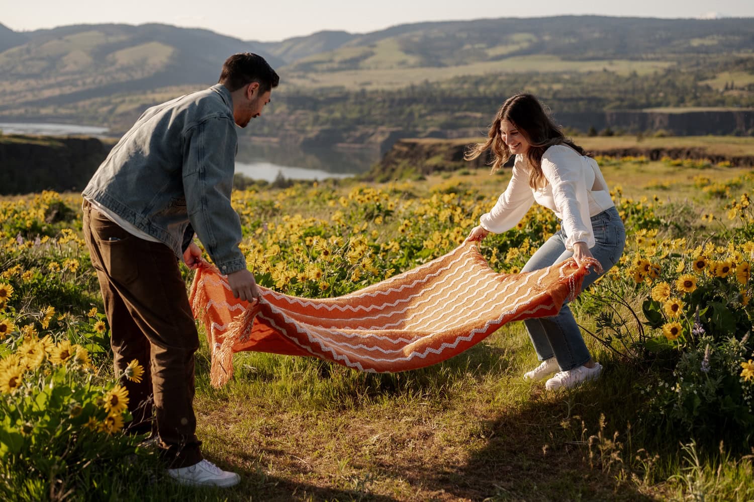 A man and woman spread a patterned blanket on green grass in a wildflower-filled field at Rowena Crest, with colorful blooms and scenic hills visible in the background.
