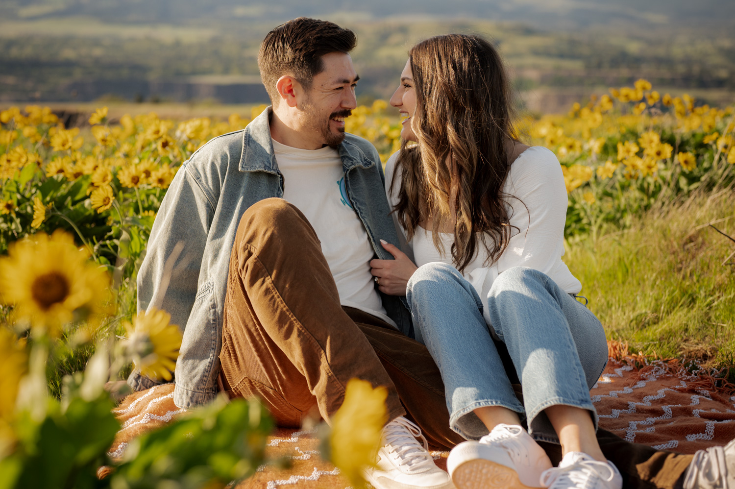 A smiling couple sits on a blanket in a sunny field of wildflowers at Rowena Crest, looking at each other.