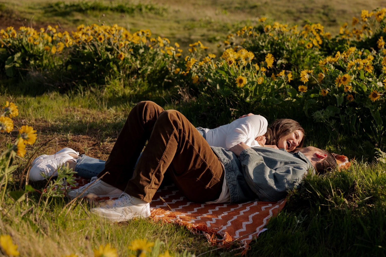 Two people lying side by side on a blanket in a field of blooming wildflowers, smiling and looking at each other with sunlight filtering through the flowers.