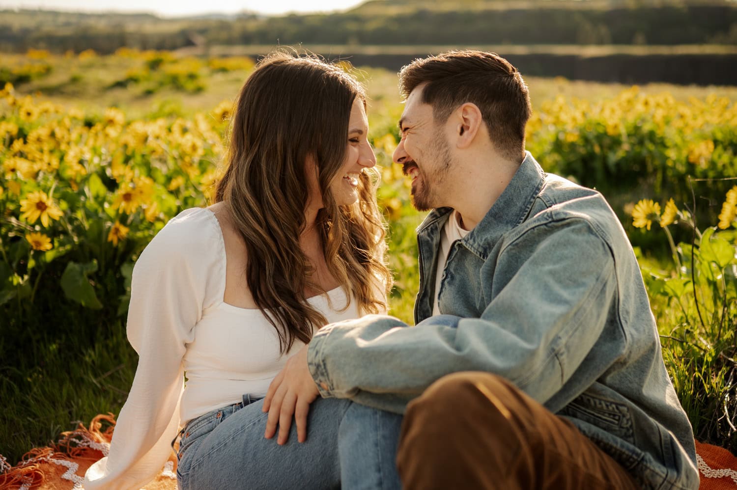 A couple sits close together in a sunny field of yellow wildflowers at Rowena Crest, smiling and looking at each other during their engagement session.