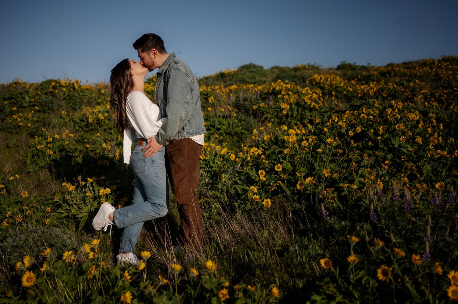 A couple embraces and smiles in a sunny field of yellow wildflowers at Rowena Crest, with a clear blue sky overhead.