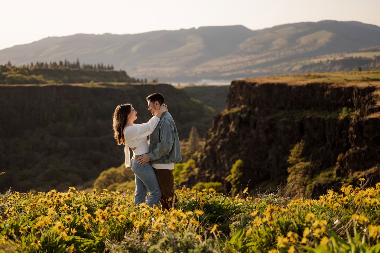 A couple embraces in a field of wildflowers at Rowena Crest with mountains visible in the background during their engagement session.