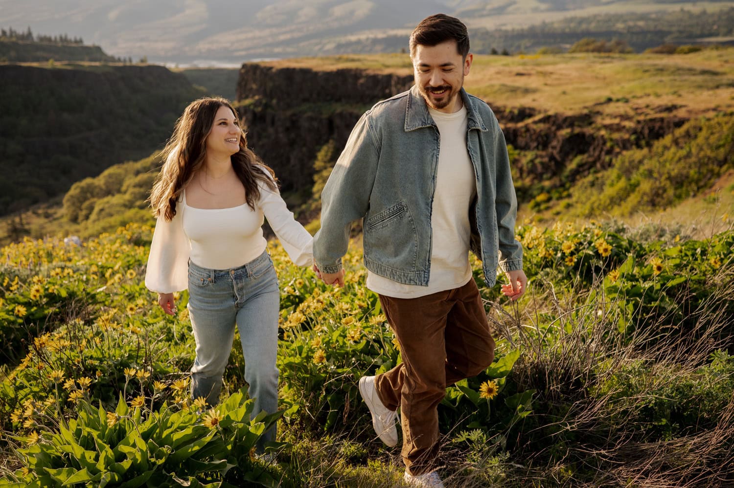 A couple holds hands while walking through a field of wildflowers, with the rolling hills of Rowena Crest visible in the background during their engagement session.