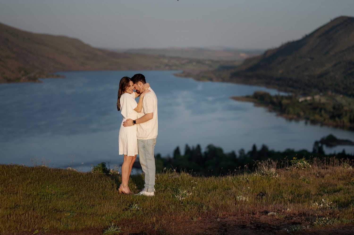 A couple embraces on a grassy hill dotted with wildflowers, overlooking a wide river and distant mountains under a clear sky at Rowena Crest.