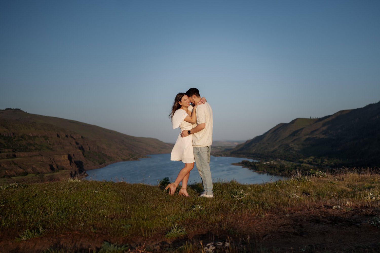 A couple embraces on a grassy hill with wildflowers, overlooking a river and mountains at sunset during their engagement session at Rowena Crest.