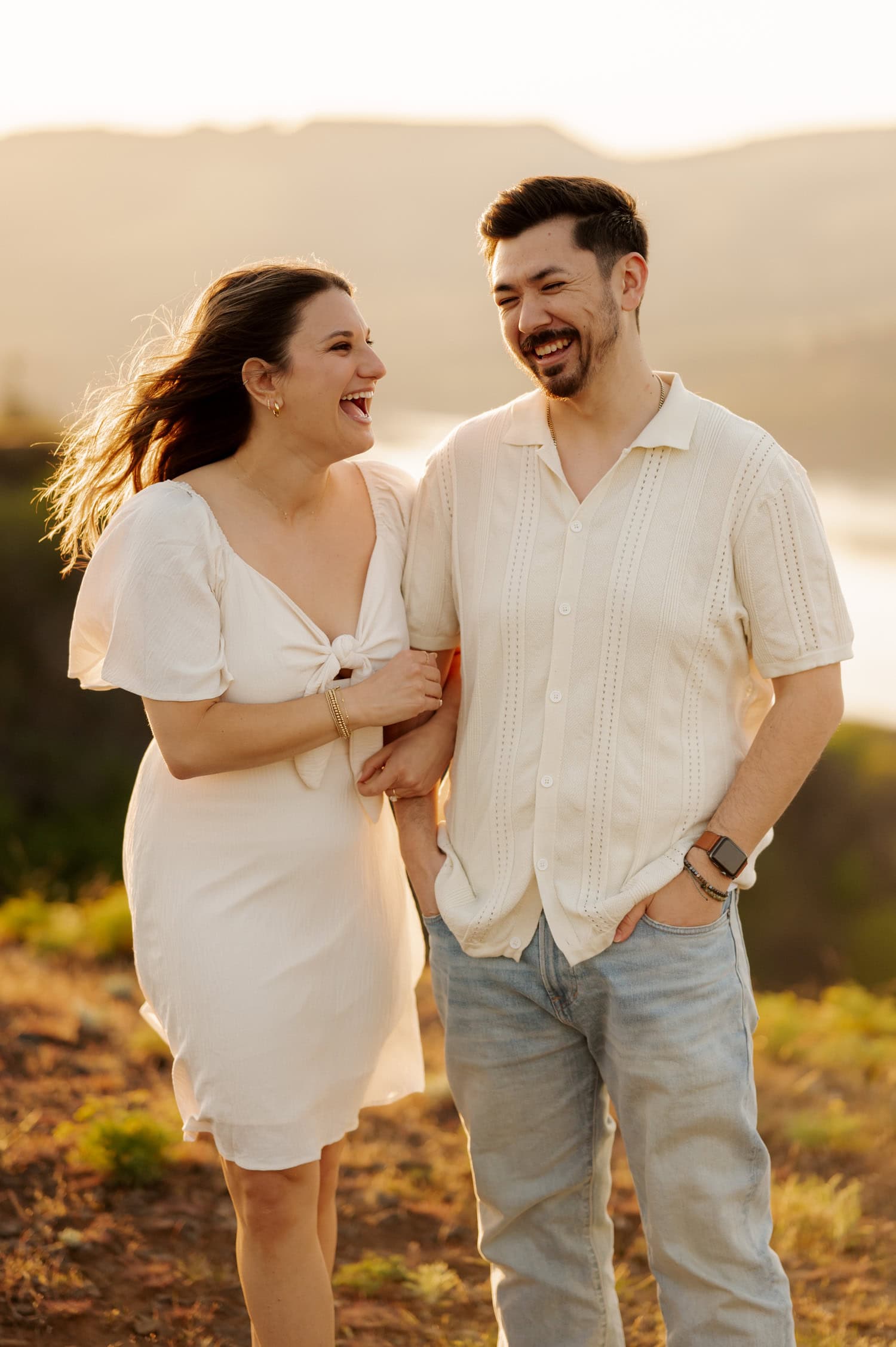 A smiling couple in light clothing laugh together during their engagement session at sunset, standing among wildflowers with hills and water visible in the background at Rowena Crest.