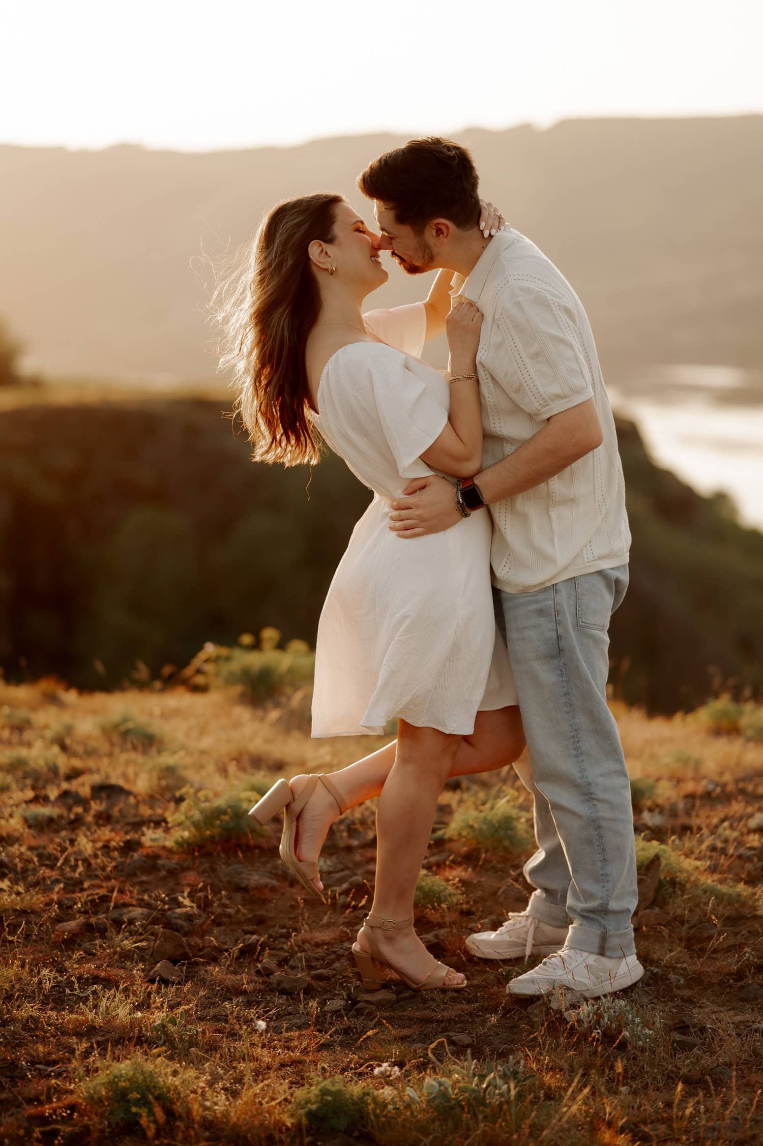 A couple embraces and smiles at sunset on a grassy hill with wildflowers, with Rowena Crest and a scenic valley visible in the background.