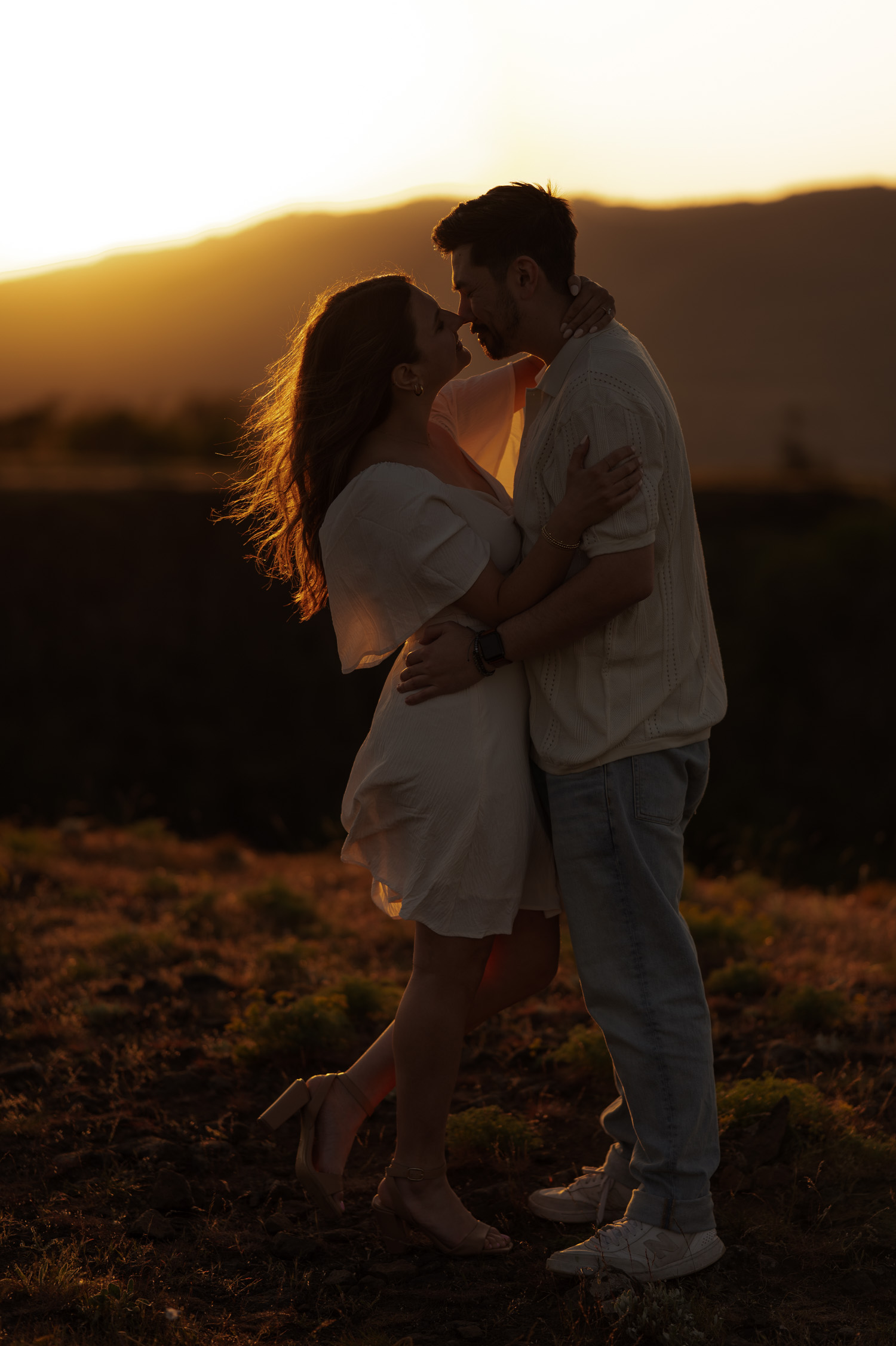 A couple embraces closely at sunset, standing outdoors among wildflowers with dramatic mountains in the background at Rowena Crest.