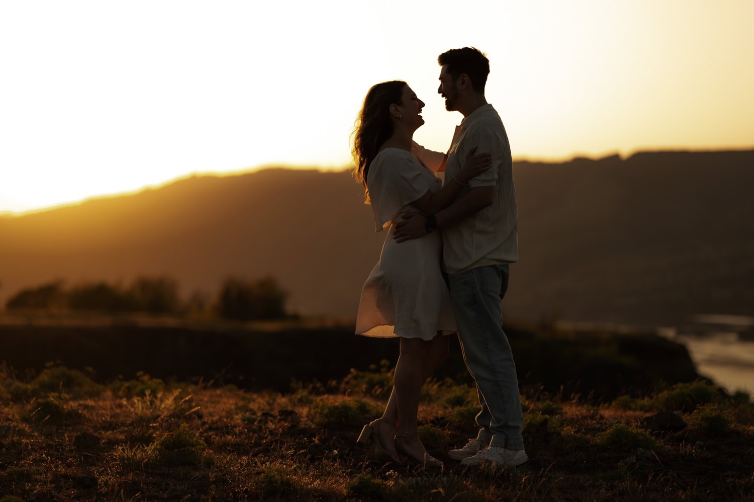A couple embraces and smiles at sunset on a grassy hill with wildflowers, mountains in the background, during their engagement session at Rowena Crest.