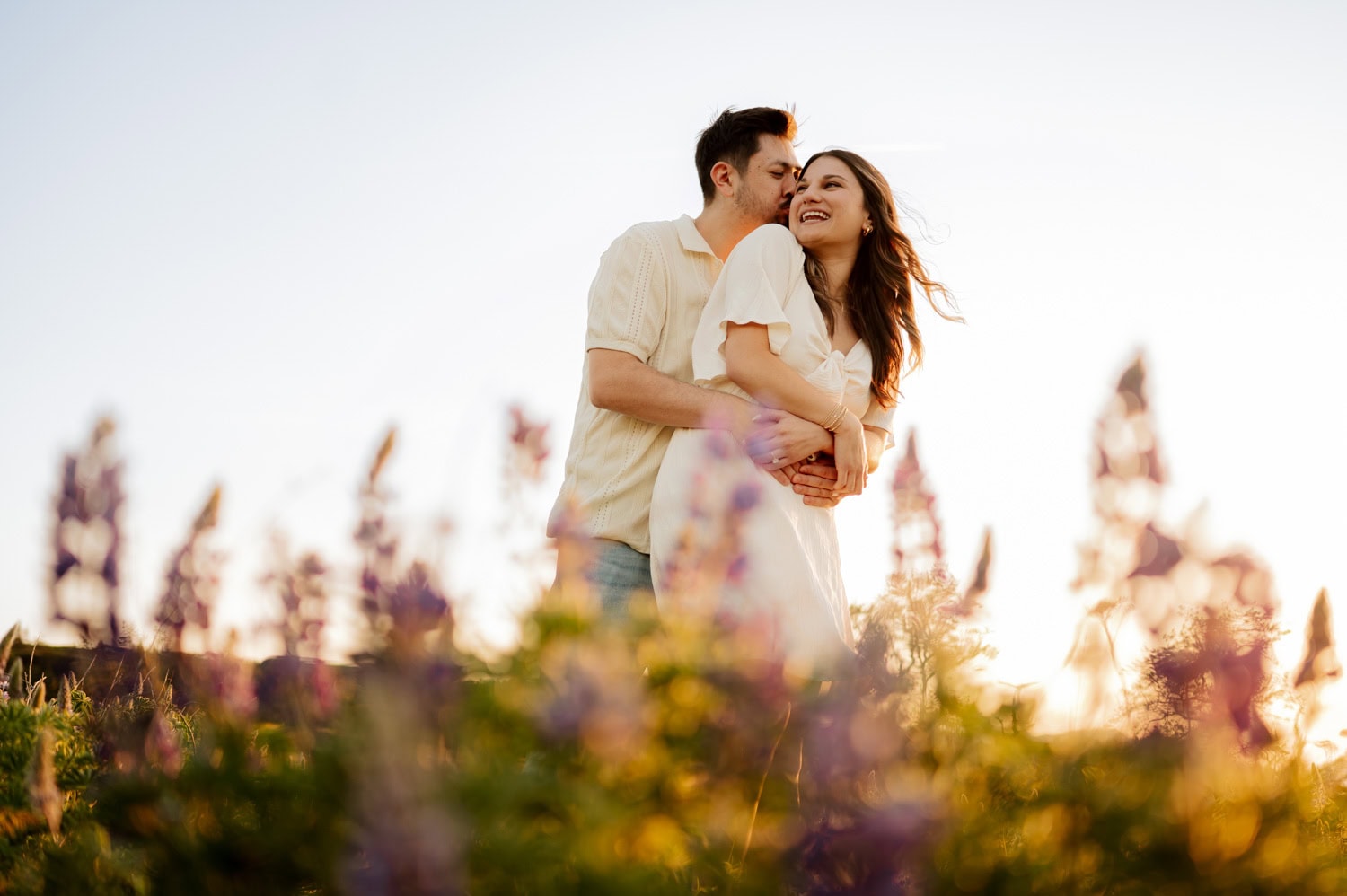 A couple hugs and smiles in a field of wildflowers at sunset, with soft sunlight glowing over Rowena Crest in the background.