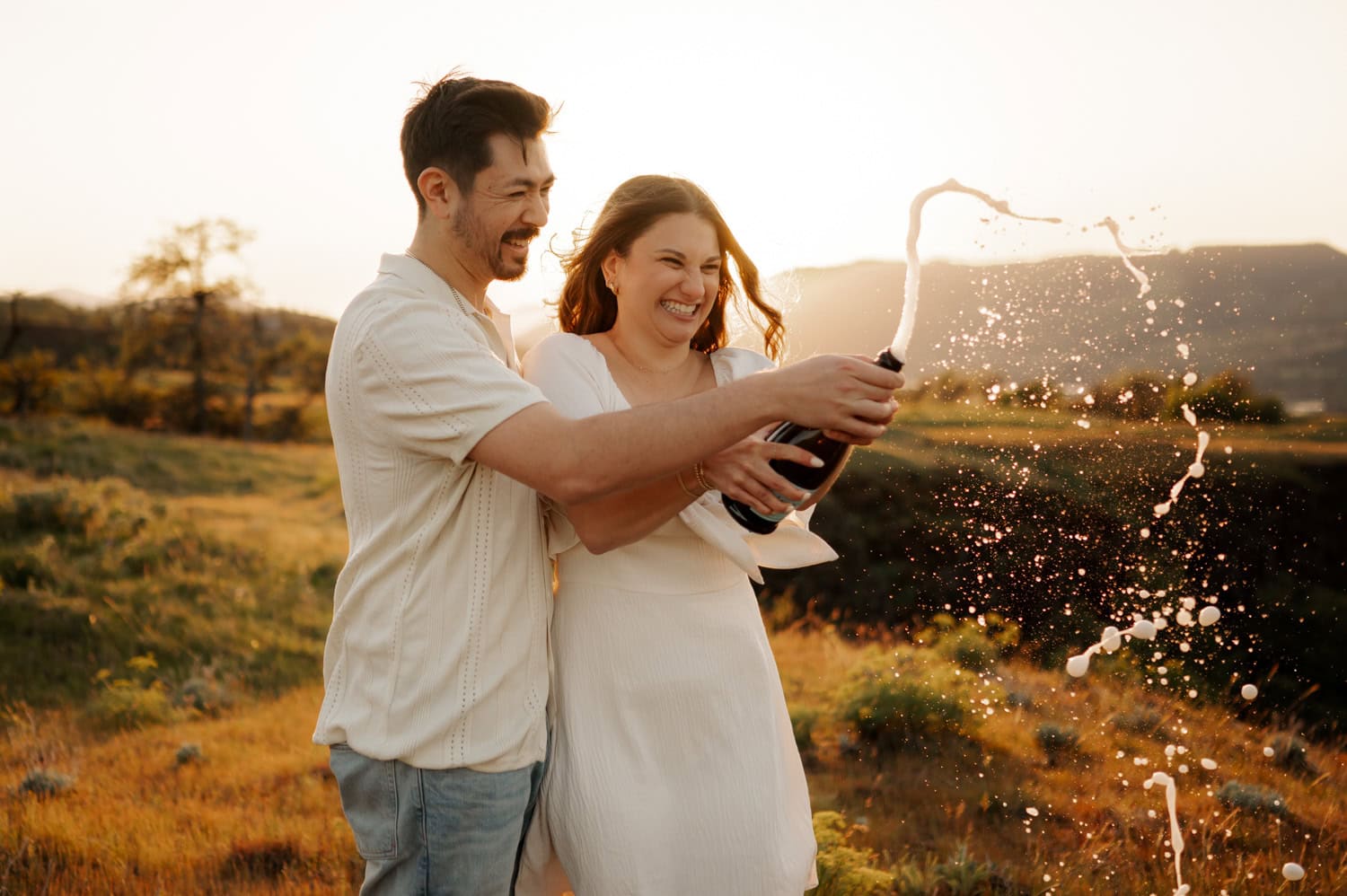 A smiling couple in light clothing pops a champagne bottle outdoors at sunset, with bubbles and foam spraying amid wildflowers during their engagement session at Rowena Crest.