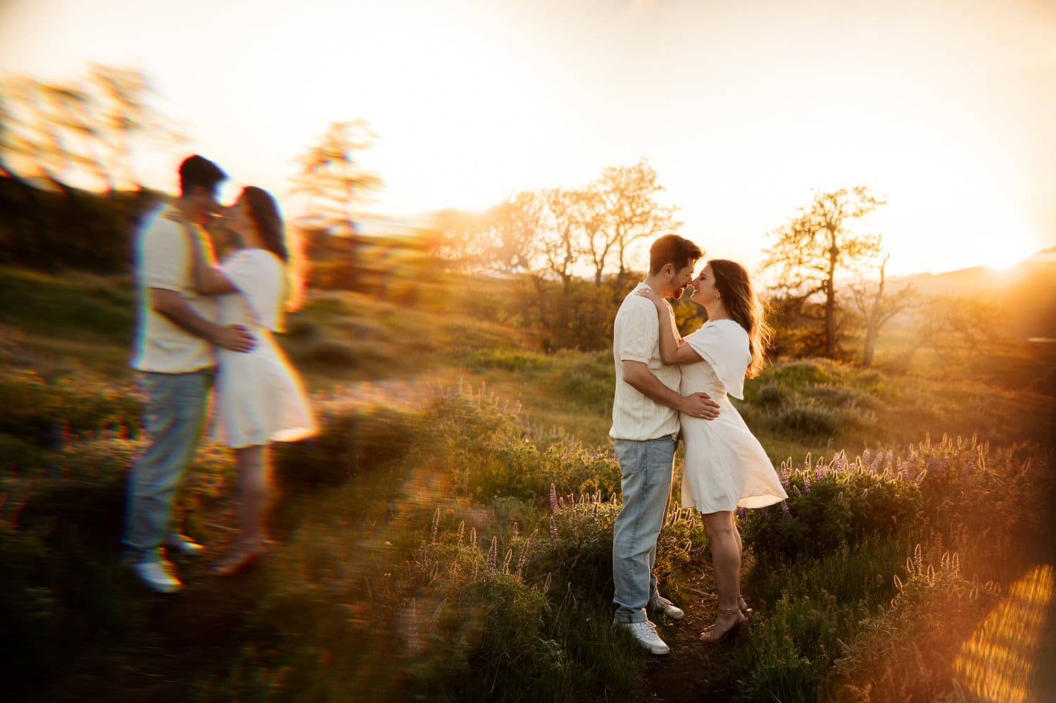 A couple kisses outdoors at sunset among wildflowers, with a blurred prism reflection of them standing together nearby.