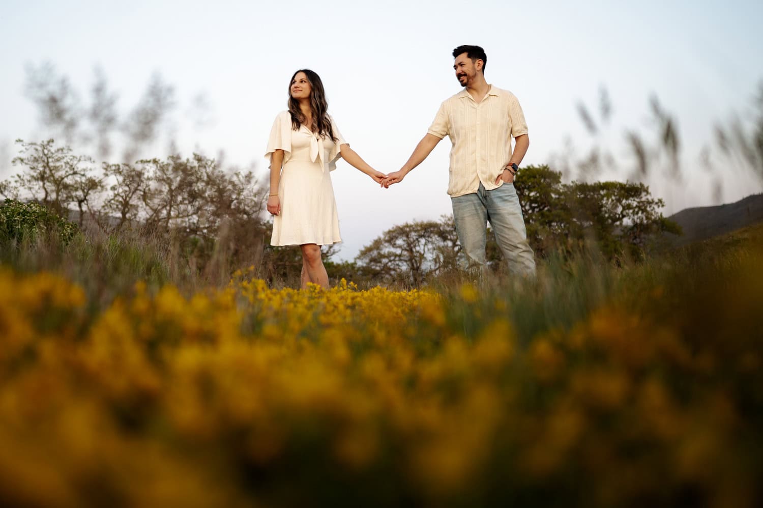 A couple holds hands in a field of wildflowers at Rowena Crest, both dressed in light-colored clothes, with trees visible in the background.