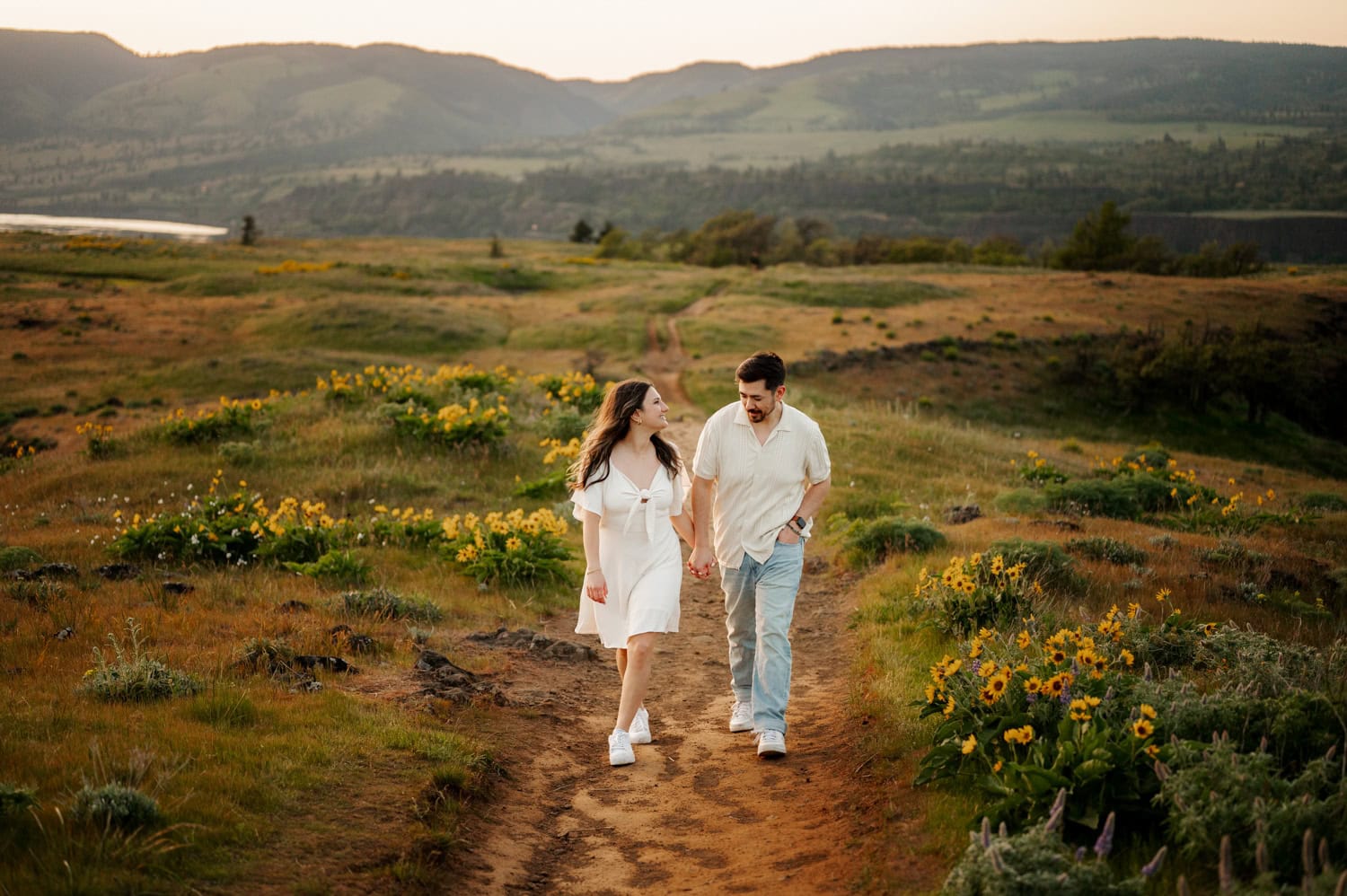 A couple walks hand in hand along a grassy path bordered by wildflowers at Rowena Crest, with rolling hills rising in the background.
