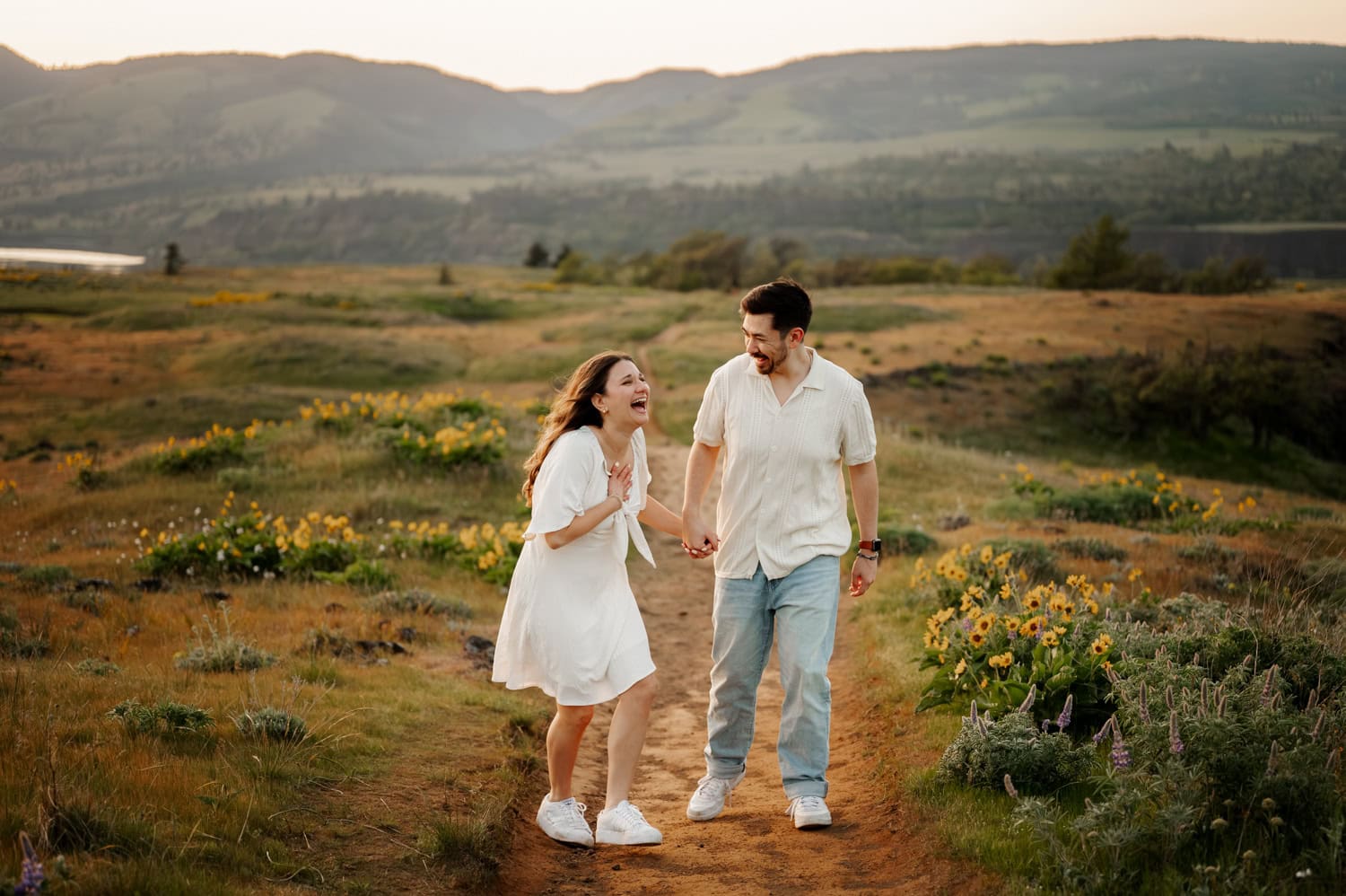 A couple laughing and holding hands while walking along a wildflower-lined path at Rowena Crest, with mountains visible in the background.